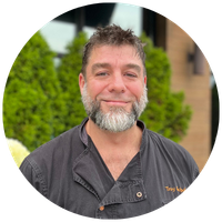 Portrait of a smiling man with a gray beard wearing a dark work shirt, outdoors with greenery behind