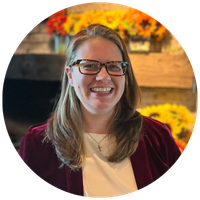 Smiling woman with glasses, shoulder-length light brown hair, wearing a white top and burgundy cardigan, in front of a colorful flower display.